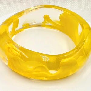 Może przedstawiać: Gruba, okrągła bransoletka typu bangle wykonana z przezroczystej żywicy z wirującymi, organicznymi wzorami z jaskrawożółtego pigmentu zatopionego w materiale. Bransoletka ma gładką, polerowaną powierzchnię oraz zaokrąglony, masywny profil.