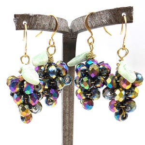 May include: A pair of drop earrings featuring clusters of iridescent, multi-coloured faceted beads shaped like grapes. Each earring includes a small, pale green leaf charm attached to a gold-toned wire hook for pierced ears.