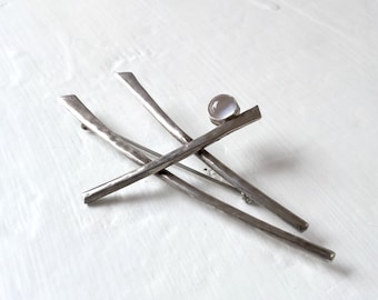 May include: A silver-toned brooch with an abstract design. The brooch features several intersecting, elongated bars with a textured finish. A small, round, light-colored stone is set on one of the bars. The brooch is displayed on a white surface.