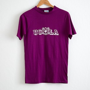 Könnte beinhalten: Ein lila T-Shirt mit einem weißen Aufdruck eines Bären und dem Text "UCLA" auf der Vorderseite.