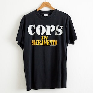 Pode incluir: Camiseta preta com texto branco que diz "COPS IN SACRAMENTO".