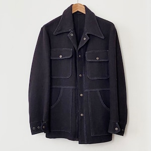 Puede incluir: Chaqueta azul marino oscuro con cuatro bolsillos delanteros y cuello. La chaqueta tiene costuras blancas visibles y botones plateados. Cuelga de una percha de madera sobre un fondo blanco.
