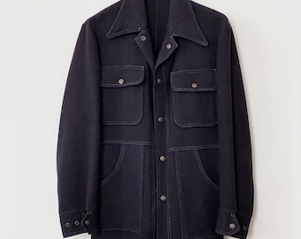 May include: A dark navy blue jacket with four front pockets and a collared neckline. The jacket has visible white stitching and silver buttons. It is hanging on a wooden hanger against a white background.