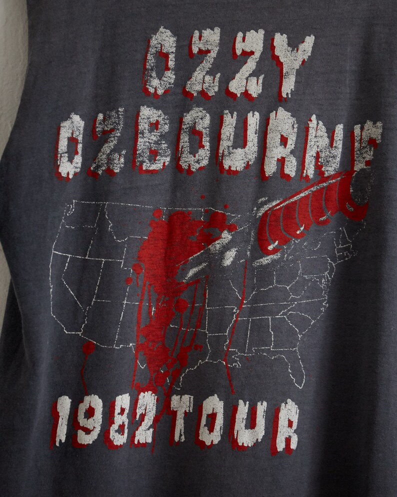 Puede incluir: Una camiseta gris sin mangas con un gr&aacute;fico blanco de la gira de Ozzy Osbourne de 1982. El gr&aacute;fico presenta un cuchillo ensangrentado y un mapa de los Estados Unidos.