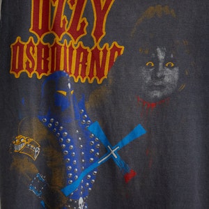 Puede incluir: Una camiseta negra con un estampado gr&aacute;fico de una figura con un traje azul y negro que sostiene una cruz azul. El texto "Ozzy Osbourne" est&aacute; impreso en letras rojas y amarillas en la camiseta.
