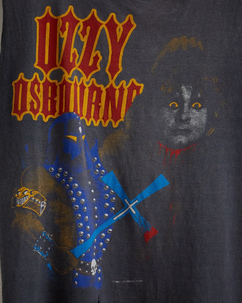 Puede incluir: Una camiseta negra con un estampado gr&aacute;fico de una figura con un traje azul y negro que sostiene una cruz azul. El texto "Ozzy Osbourne" est&aacute; impreso en letras rojas y amarillas en la camiseta.
