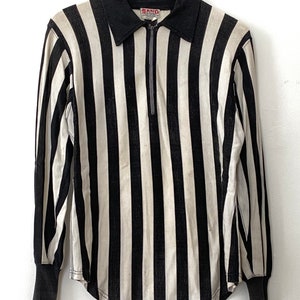 Puede incluir: Camisa de manga larga a rayas blancas y negras con cierre de cremallera y la palabra "SANO" en la parte delantera.