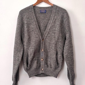 May include: A gray cardigan sweater with a V-neck, button closure, and two front pockets. The sweater is made of a textured knit fabric and hangs on a wooden hanger. A small label is visible near the neckline.