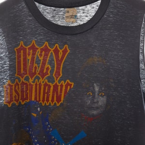 Puede incluir: Una camiseta gris sin mangas con un estampado descolorido de Ozzy Osbourne. El estampado presenta el nombre de la banda en una fuente estilizada con un contorno amarillo y letras rojas. Tambi&eacute;n se incluye una imagen en blanco y negro de Ozzy Osbourne en el estampado.