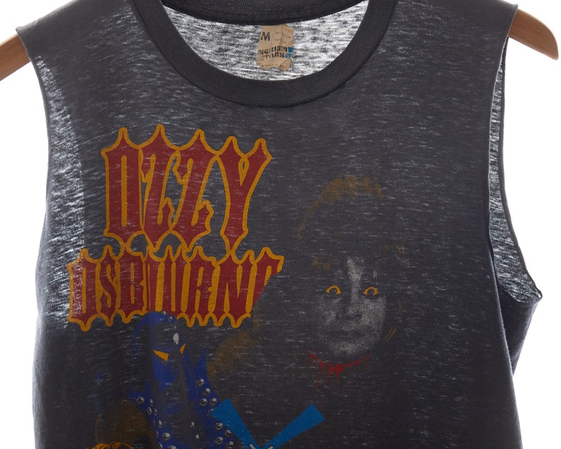 Puede incluir: Una camiseta gris sin mangas con un estampado descolorido de Ozzy Osbourne. El estampado presenta el nombre de la banda en una fuente estilizada con un contorno amarillo y letras rojas. Tambi&eacute;n se incluye una imagen en blanco y negro de Ozzy Osbourne en el estampado.