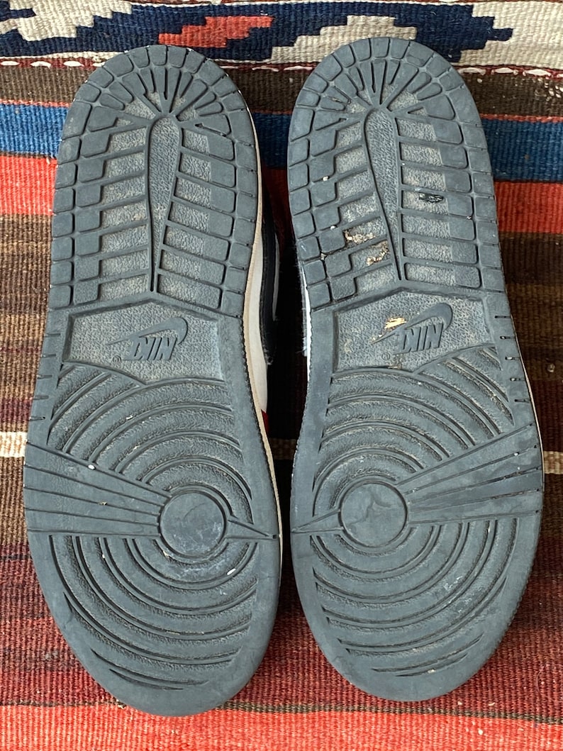 May include: A pair of black and white sneakers with red accents. The sneakers have a black sole with a circular pattern and the Nike logo on the back.