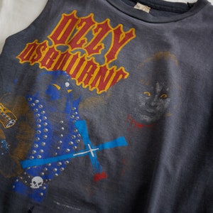 Puede incluir: Una camiseta sin mangas de color gris oscuro con un estampado gr&aacute;fico de una banda llamada "Ozzy Osbourne". El gr&aacute;fico presenta a una persona con una chaqueta azul y plateada tachonada y un gato con ojos rojos.