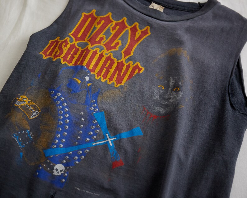 Puede incluir: Una camiseta sin mangas de color gris oscuro con un estampado gr&aacute;fico de una banda llamada "Ozzy Osbourne". El gr&aacute;fico presenta a una persona con una chaqueta azul y plateada tachonada y un gato con ojos rojos.