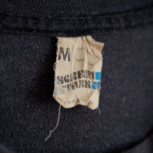 Puede incluir: Un primer plano de una etiqueta de camiseta negra con fondo blanco. La etiqueta dice "M" y "Screamer T-Shirts" en texto negro. La etiqueta tambi&eacute;n dice "100% Algod&oacute;n" y "Hecho en EE. UU."