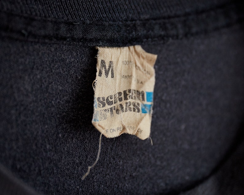 Puede incluir: Un primer plano de una etiqueta de camiseta negra con fondo blanco. La etiqueta dice "M" y "Screamer T-Shirts" en texto negro. La etiqueta tambi&eacute;n dice "100% Algod&oacute;n" y "Hecho en EE. UU."