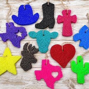 May include: A collection of twelve colorful, textured aroma bead ornaments in various shapes including a blue cowboy hat, black boot, pink cross, purple Texas state, teal cow head, blue fleur-de-lis, yellow star, brown deer head, red heart, pink Texas state, and green cactus.