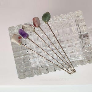 Könnte beinhalten: Fünf Cocktailspieße aus Metall mit gedrehten Stielen, jeweils gekrönt von einem einzigartigen polierten Edelstein in Weiß, Violett, Transparent, Rosa und Grün, die diagonal auf einer klaren, rechteckigen Glaskaraffe mit Gittermuster liegen.