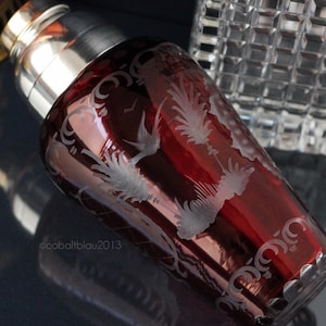 Puede incluir: Una coctelera de vidrio roja con tapa plateada y diseños florales y de pájaros grabados.