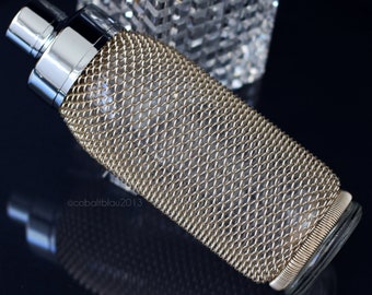 May include: A gold and silver bottle with a textured, mesh-like design. The bottle has a silver cap and a clear glass base.