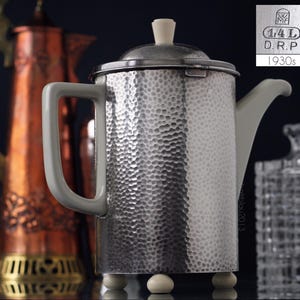 Peut inclure: Cafetière en argent martelé avec une poignée et un couvercle blancs. La cafetière a une capacité de 1,4 L et est marquée "D.R.P. 1930s".
