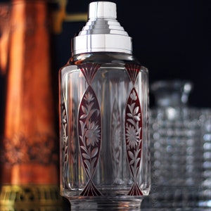 Puede incluir: Un coctelera de cristal transparente con tapa plateada y diseño floral rojo y blanco. La coctelera tiene una superficie texturizada y una base redonda.