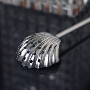 Puede incluir: Una cuchara de bar de acero inoxidable con un colador en forma de concha. El colador tiene múltiples agujeros pequeños. La cuchara está sobre una superficie oscura y reflectante.