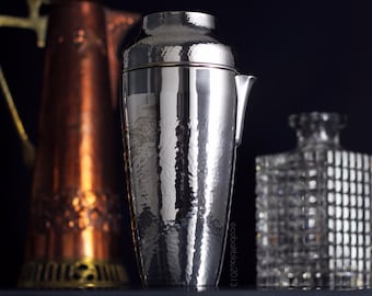 Puede incluir: Un coctelera de plata con acabado martillado. La coctelera tiene una parte superior redondeada y un pico en el lateral. La coctelera está sobre una superficie negra.