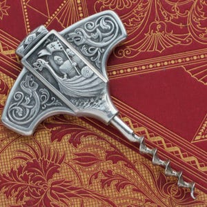Puede incluir: Un abridor de botellas de vino plateado con un diseño detallado de barco vikingo. El abridor presenta un sacacorchos y un mango decorativo en forma de martillo. El mango tiene intrincadas tallas y un panel rectangular que representa un barco vikingo.