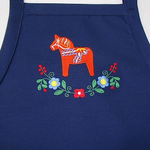 May include: A navy blue apron featuring an embroidered orange Dala horse design. The horse is surrounded by a floral wreath with blue, red, and green accents. The apron has a simple, functional design.