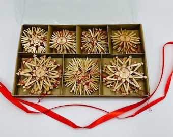 May include: A set of six star-shaped ornaments crafted from natural straw, arranged in a partitioned cardboard box. Each ornament features intricate geometric patterns bound with thin red thread, and a long red satin ribbon extends from the box.