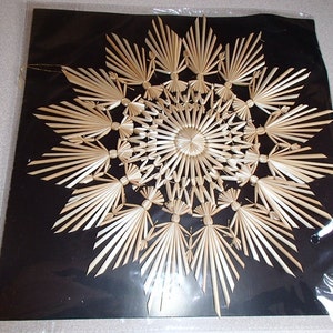May include: A decorative straw ornament in a starburst design. The natural-colored straw is arranged on a black background. The ornament is encased in clear plastic and has a gold string for hanging.