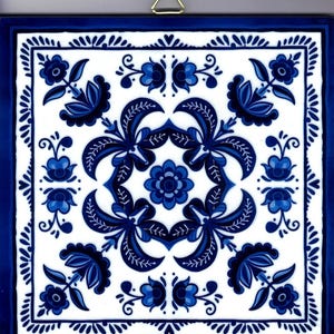 May include: Square ceramic tile with a white background and a blue floral design. The design features stylized flowers and leaves in varying shades of blue, framed by a dark blue border. A gold-colored metal hanger is attached to the top.