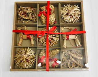 May include: A set of nine straw ornaments in a divided cardboard box, tied with a red ribbon. The ornaments include starbursts, pine cones, and a straw horse. The box is presented on a white surface.
