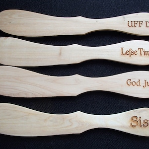 May include: Four wooden lefse turners with engraved text. The text includes "UFF DA", "Lefse Time", "God Jul", and "Sisu". The turners are light-colored wood with a smooth finish, and a curved handle. The background is a dark, solid color.