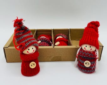 May include: Red and gray knitted Christmas ornaments in the shape of dolls. The dolls have smiling faces, red hats, and red and gray striped sweaters. One doll is outside the cardboard box, and three are inside.