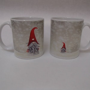 May include: Two white ceramic mugs with a winter gnome design. The gnomes wear red hats and have gray beards. The mugs have a winter scene background with falling snow.