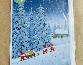 May include: A rectangular advent calendar featuring a snowy winter forest scene with two gnomes pulling a sled filled with gifts, a small house, and a rabbit. The design includes numbered perforated doors and a green circular Bio-Glitter label.
