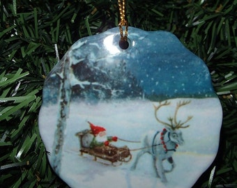 May include: A ceramic Christmas ornament with a winter scene featuring a reindeer pulling a sleigh with a small figure in a red hat. The ornament has a gold string for hanging and is set against a backdrop of green Christmas tree needles.