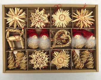 May include: A set of Christmas ornaments in a cardboard box. The ornaments include straw stars, reindeer, gnomes with red hats, and heart-shaped decorations. The ornaments are made of natural straw and red string.