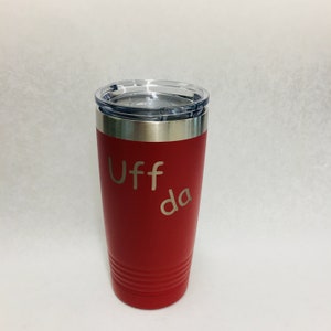 May include: A red insulated tumbler with a silver rim and clear lid. The tumbler has the word "Uff da" etched in a light color on the front. The tumbler is cylindrical with horizontal grooves near the bottom.