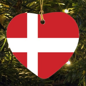 May include: Heart-shaped Christmas ornament featuring the flag of Denmark. The ornament has a red background with a white Scandinavian cross design. A gold string is attached for hanging on a Christmas tree.