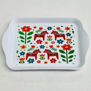 May include: White rectangular serving tray with rounded handles. The tray features a colorful pattern of red Dala horses, flowers, and green leaves. The floral design includes red, blue, and yellow flowers, creating a vibrant and cheerful aesthetic.