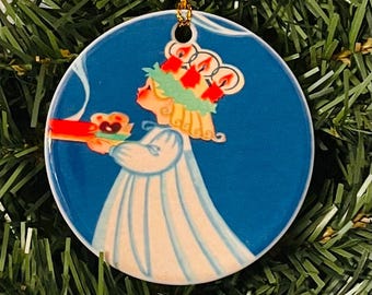 May include: Round Christmas ornament with a blue background featuring a vintage illustration of a girl in a white gown carrying a tray with a heart-shaped pastry. The girl wears a wreath of candles on her head. A gold string is attached for hanging.