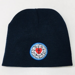 May include: A navy blue beanie hat with a circular embroidered patch. The patch features a white rose with a red heart and a black cross in the center, surrounded by a light blue border.