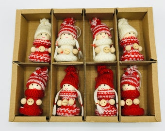May include: Eight Christmas ornaments in a cardboard box. Each ornament is a small doll with a knitted red and white outfit, a matching hat, and wooden button details. The dolls have white yarn hair and are designed for holiday decor.