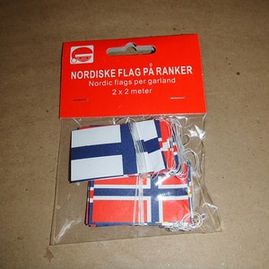 May include: A package of Nordic flag garlands. The flags are red, white, and blue, and are strung on white string. The package is red with white text that reads "Nordic flags per garland 2 x 2 meter."