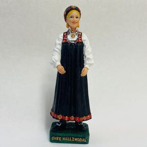 May include: A figurine of a woman in traditional Norwegian attire. She wears a black dress, white blouse, and a colorful headscarf. The base is green with the text "ØVRE HALLINGDAL". The figurine has a smiling expression and is a decorative item.