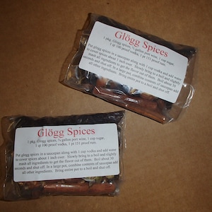 May include: Two clear plastic bags of Glögg Spices, each with a printed label. The labels list ingredients like port wine, sugar, vodka, and rum. Instructions for use are also printed on the labels. The bags contain a mix of spices.