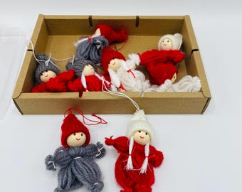May include: A collection of handmade doll ornaments in a cardboard box. The dolls have yarn bodies and hats in red, gray, and white. The dolls are designed with smiling faces and are suitable for holiday decorations.