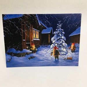 May include: A rectangular greeting card featuring a winter night scene with three gnomes in red hats. One gnome sits by a dark cabin door with a lantern, another pulls a small wooden sled, and the third stands by a snow-covered pine tree.